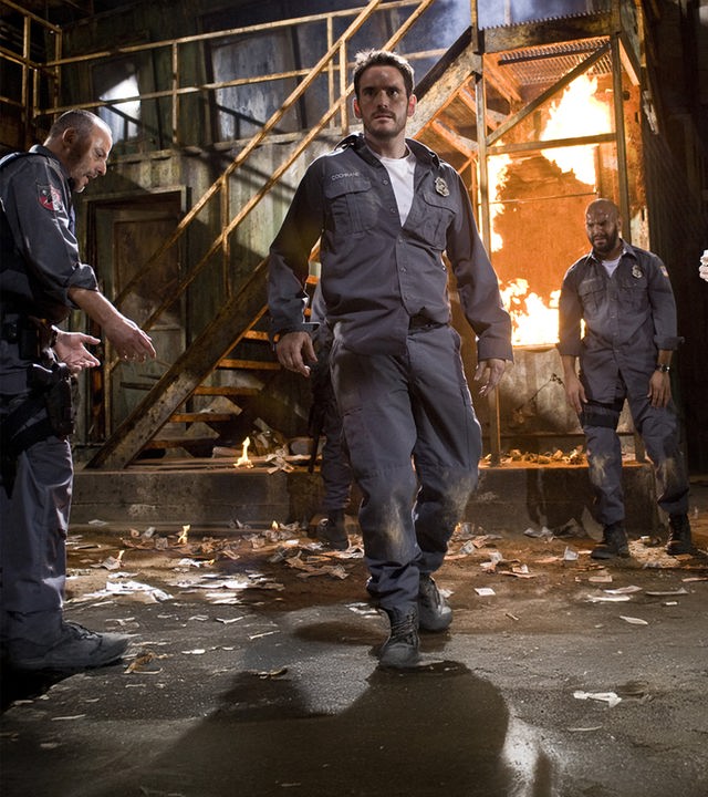 "Armored": Die uniformierten Wächter eines Geldtransports Quinn (Jean Reno), Mike Cochrane (Matt Dillon), Palmer (Amaury Nolasco) und Dobbs (Skeet Ulrich) stehen in einer Fabrikhalle vor einer brennenden Treppe und schauen angespannt zur anderen Seite des Raumes.