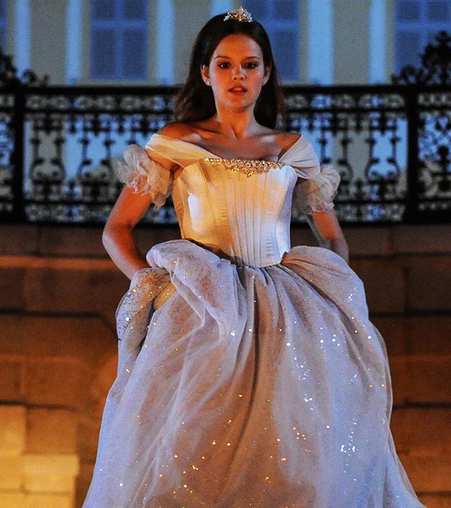 Im Hintergrund das nächtliche Schloss. Aschenputtel (Emilia Schüle), gekleidet in ein schneeweißes Kleid, hat das Kleid in den Hüften etwas angehoben, um den Ball schnell durch die geöffnete, eiserne Toreinfahrt des Schlosses zu verlassen.