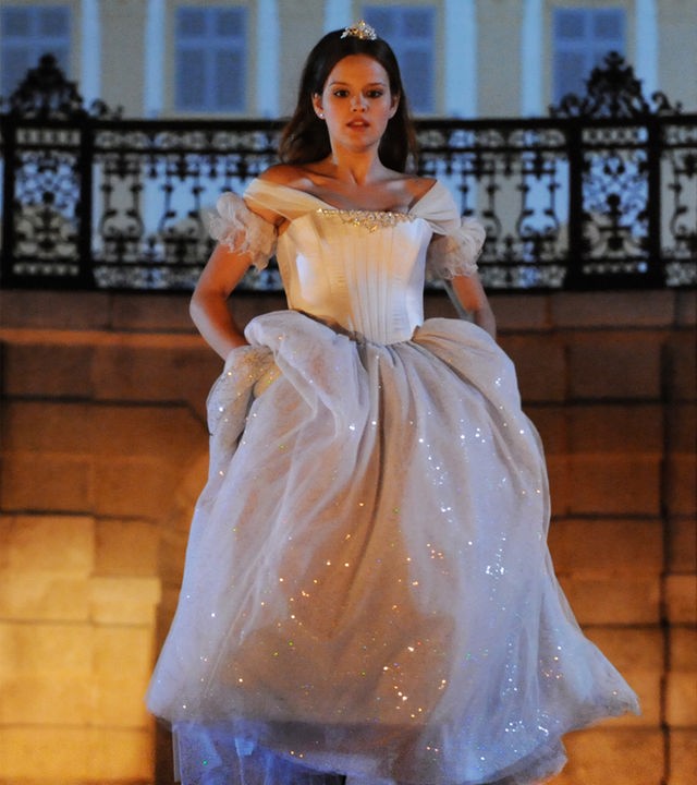 Im Hintergrund das nächtliche Schloss. Aschenputtel (Emilia Schüle), gekleidet in ein schneeweißes Kleid, hat das Kleid in den Hüften etwas angehoben, um den Ball schnell durch die geöffnete, eiserne Toreinfahrt des Schlosses zu verlassen.