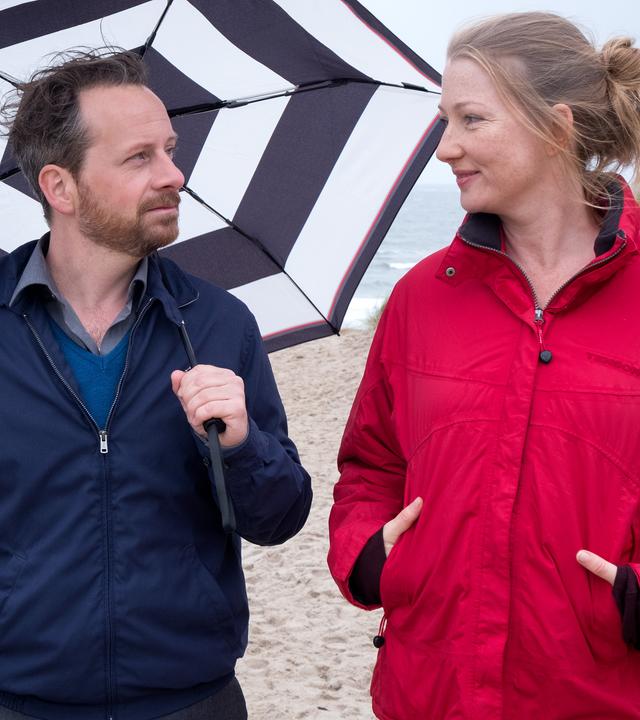 Kevin (Fabian Busch) und Bente (Katja Studt) kommen sich bei einem gemeinsamen Strandspaziergang näher. "Ausgerechnet Sylt": Kevin (Fabian Busch) und Bente (Katja Studt) spazieren gemeinsam am Strand entlang. Er hat einen Regenschirm über sich aufgespannt und schaut Bente lächelnd an, die seinen Blick erwidert. Im Hintergrund ist das Meer zu sehen.