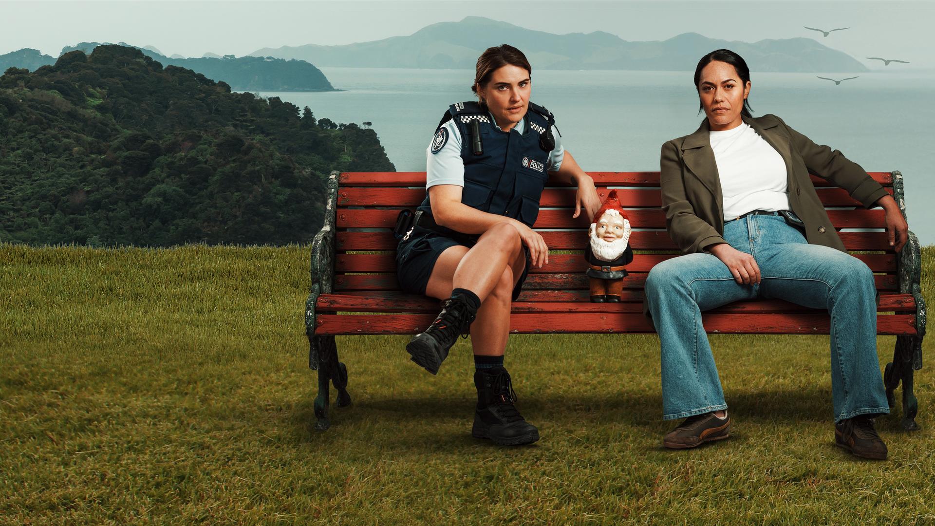Deb und Mihi, die im entlegenen Northland (Neuseeland) als Polizistinnen zusammenarbeiten, nachdem ihre Liebesbeziehung abrupt endete. Sie sitzen nebeneinander auf einer Bank, die auf einer Wiese steht. Zwischen ihnen steht ein kleiner Gartenzwerg. Im Hintergrund sieht man die bewaldete Küste und das Meer.