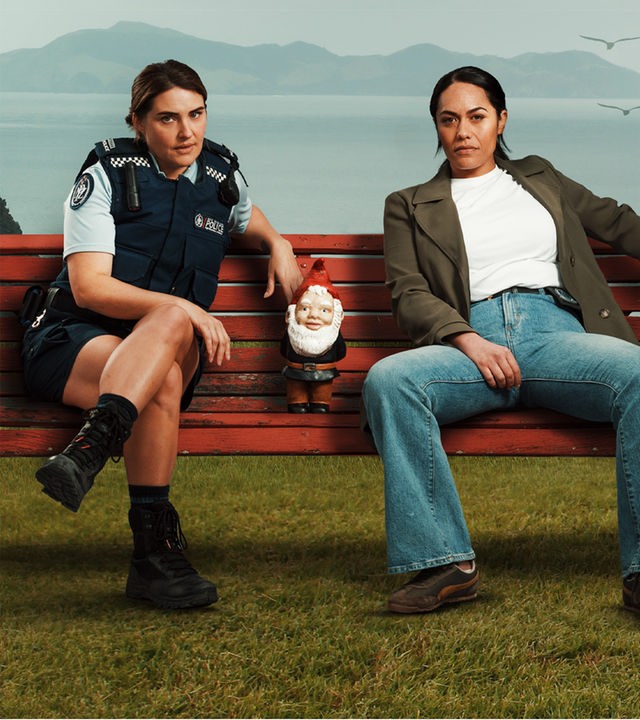 Deb und Mihi, die im entlegenen Northland (Neuseeland) als Polizistinnen zusammenarbeiten, nachdem ihre Liebesbeziehung abrupt endete. Sie sitzen nebeneinander auf einer Bank, die auf einer Wiese steht. Zwischen ihnen steht ein kleiner Gartenzwerg. Im Hintergrund sieht man die bewaldete Küste und das Meer.