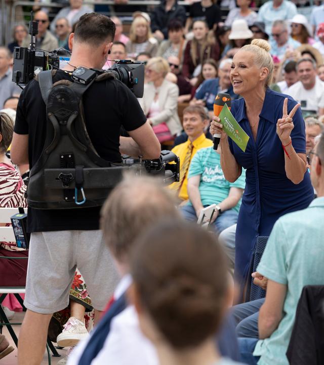 "Backstage - Fernsehgarten": Andrea "Kiwi" Kiewel steht mitten im Publikum im Fernsehgarten. Sie hält ein Mikrofon und Moderationskarten in der Hand. Sie schaut in eine Kamera, die ein Kameramann vor ihr mit sich trägt. 
