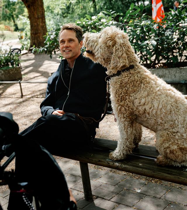 Schauspieler Matthias Schloo sitzt lächelnd auf einer Parkbank. Neben ihm auf der Bank sitzt sein großer Hund "Bowie" mit beigem-lockigem Fell