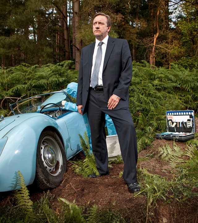 John Barnaby (Neil Dudgeon) steht im Anzug in einem Waldstück neben einem hellblauen Oldtimer-Cabriolet, das abseits des Weges im Gebüsch gelandet ist. Rechts auf dem Boden ist ein Koffer mit Werkzeugen angeschnitten zu sehen, während im Hintergrund eine Person in einem blauen Schutzanzug am Wagen arbeitet.