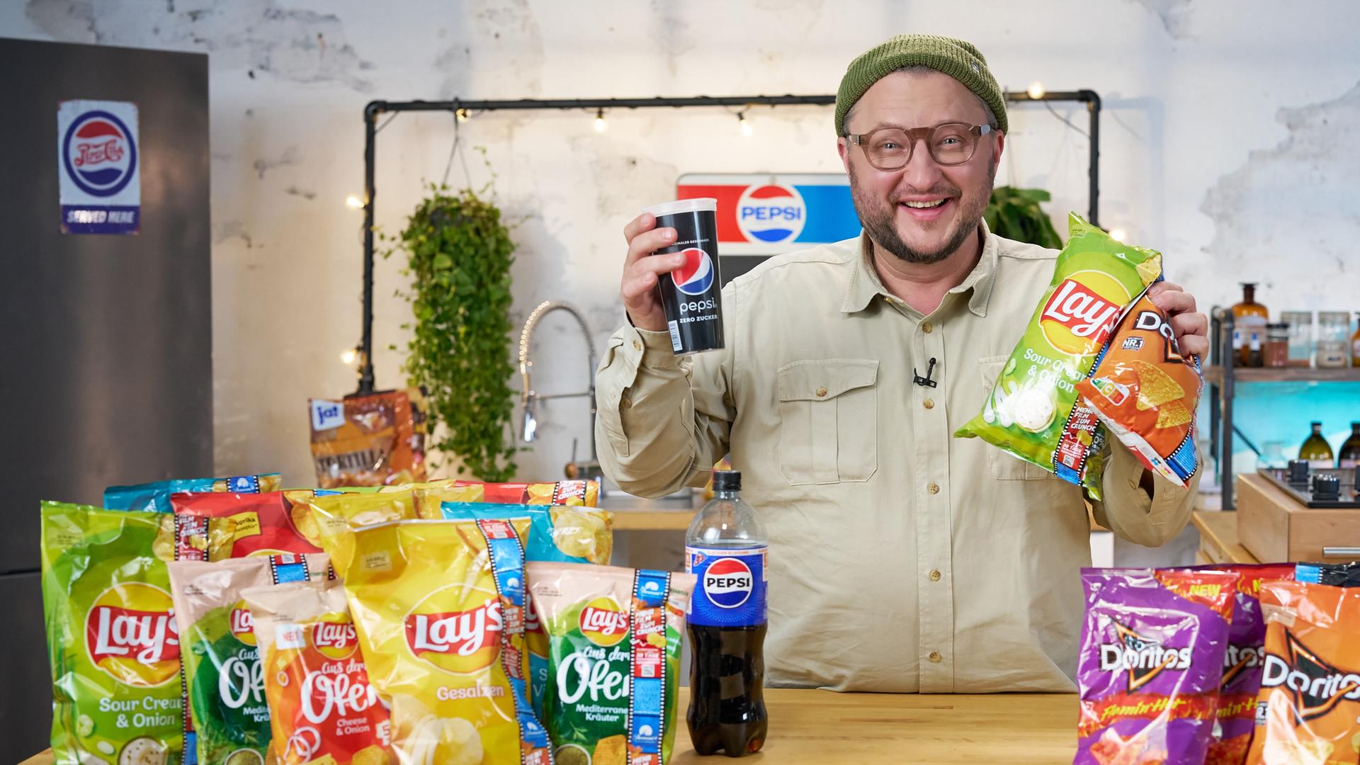 Produktentwickler Sebastian Lege hält einen Becher mit dem Aufdruck "Pepsi" und zwei Chips-Tüten in der Hand, darauf steht "Lays" und "Doritos". Auf dem Tisch vor Sebastian Lege liegen diverse Chipstüten unterschiedlicher Marken, die zum Konzern PepsiCo gehören.