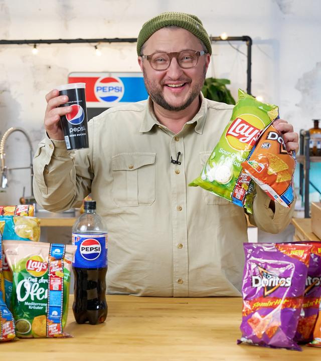 Produktentwickler Sebastian Lege hält einen Becher mit dem Aufdruck "Pepsi" und zwei Chips-Tüten in der Hand, darauf steht "Lays" und "Doritos". Auf dem Tisch vor Sebastian Lege liegen diverse Chipstüten unterschiedlicher Marken, die zum Konzern PepsiCo gehören.
