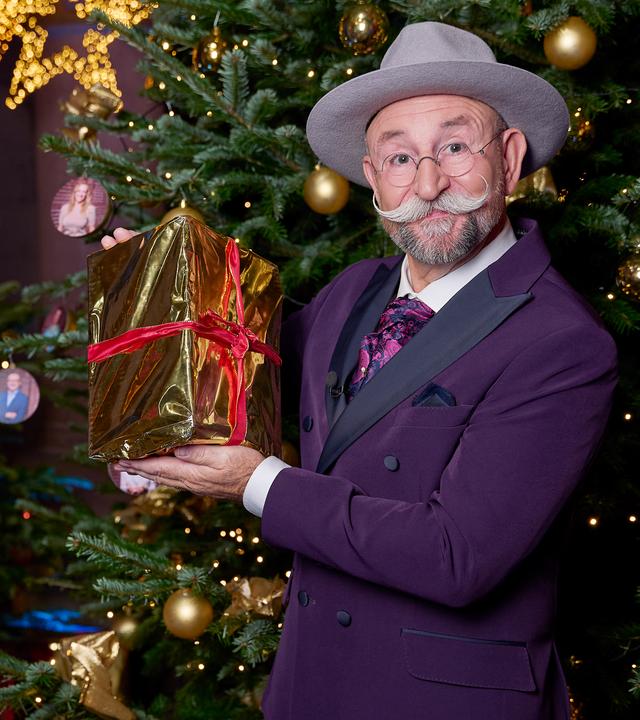 Horst Lichter steht vor geschmückten Weihnachtsbäumen und hält die Händlerkarte in den Händen