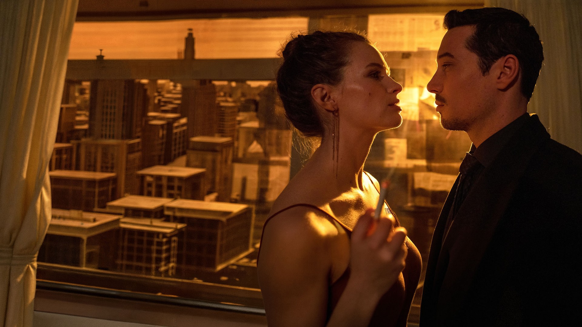 "Blindspot": Anara van Veeren (Marlene Tanczik) und Max van Veeren (Klaus Steinbacher) stehen sich eng gegenüber und schauen sich gegenseitig mit ernster Miene an. Anara hält in ihrer rechten Hand eine brennende Zigarette. Durch das mit Zeitungspapier verdunkelte Fenster scheint gelbes Licht.