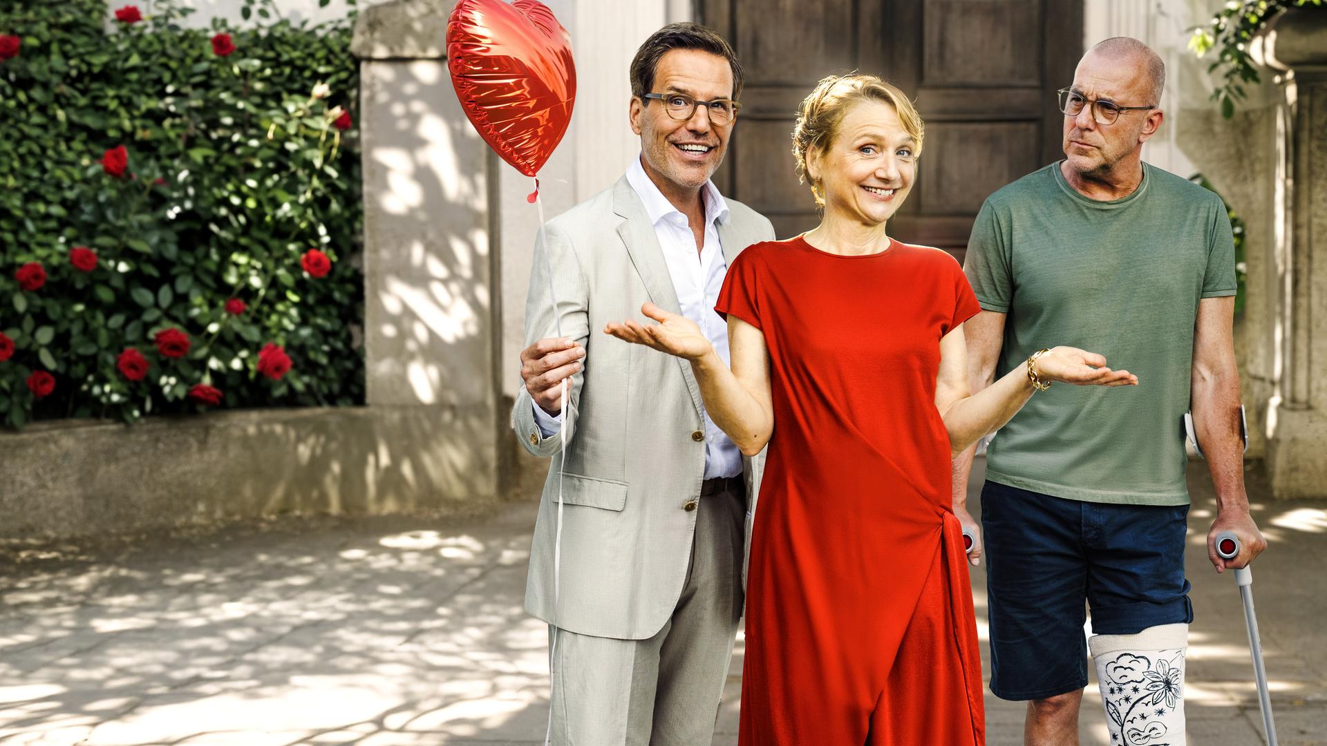 "Bloß nicht Liebe": Vor einem Münchner Wohnhaus stehen Benno Martius (Tim Bergmann), Viola Berger (Christina Große) und Dr. Justus Förster (Heino Ferch) nebeneinander. Benno Martius, in einem eleganten Anzug, hält lachend einen roten Herzluftballon in der Hand. Neben ihm steht, mit Gipsbein und auf Krücken Dr. Förster, der Martius kritisch mustert. Zwischen den beiden Männern befindet sich in einem roten Kleid Viola Berger. Sie lacht, hält aber die Hände unentschlossen in die Luft.