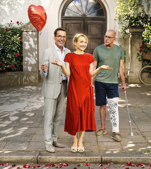 "Bloß nicht Liebe": Vor einem Münchner Wohnhaus stehen Benno Martius (Tim Bergmann), Viola Berger (Christina Große) und Dr. Justus Förster (Heino Ferch) nebeneinander. Benno Martius, in einem eleganten Anzug, hält lachend einen roten Herzluftballon in der Hand. Neben ihm steht, mit Gipsbein und auf Krücken Dr. Förster, der Martius kritisch mustert. Zwischen den beiden Männern befindet sich in einem roten Kleid Viola Berger. Sie lacht, hält aber die Hände unentschlossen in die Luft.