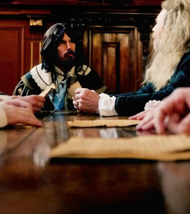 "Charles I. - Machtkampf in England: Der Bürgerkrieg": Spielszene: In einem holzgetäfelten Saal sitzen fünf Männer an einem großen, dunklen Tisch, der teilweise mit Papieren bedeckt ist. Der Mann in der Mitte wird auf jeder Seite von zwei Personen flankiert, die auf den Mann schauen. 