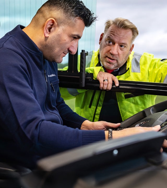 Ein Mann sitzt am Steuer eines Linienbusses und schaut auf das Lenkrad. Draußen steht der Fahrlehrer und beugt sich zum Fenster hinein, während er ihm das Buscockpit erklärt. Links unten befindet sich ein weißes Titellogo in Form eines Lenkrads mit dem Schriftzug: „Das fahrende Klassenzimmer“. 