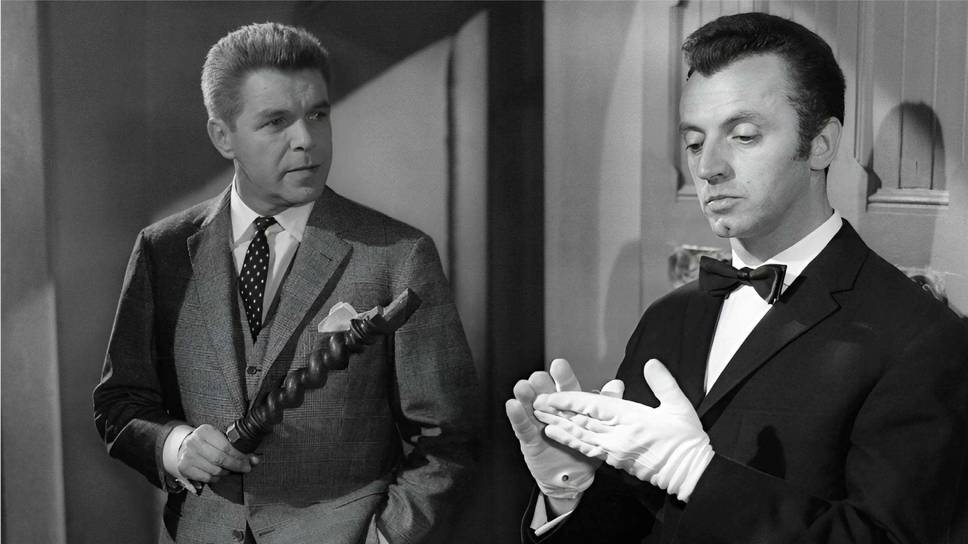 Der Anwalt Frank Tanner (Heinz Drache, links) hält eine Art hölzernes Möbelstück in der Hand, während er mit dem Butler Bonwit (Eddy Arendt) spricht, der sich gerade die weißen Handschuhe glattstreicht.