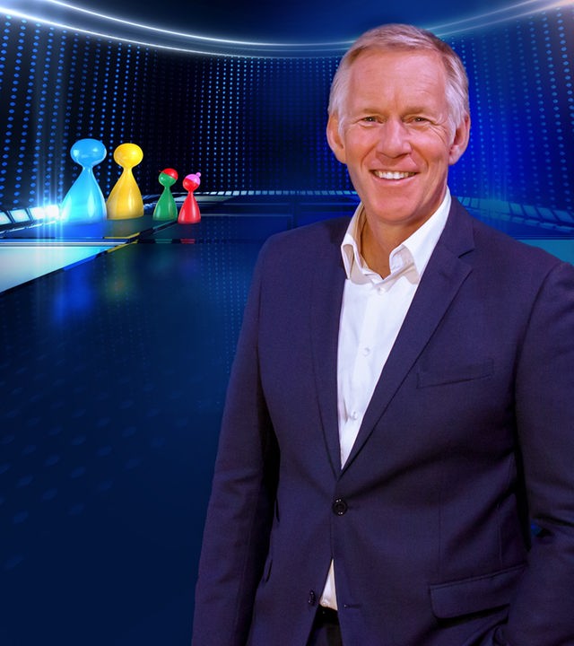 Johannes B. Kerner steht vor LED-Wand mit "Spiel beginnt!"-Logo im Studio und lächelt in die Kamera.