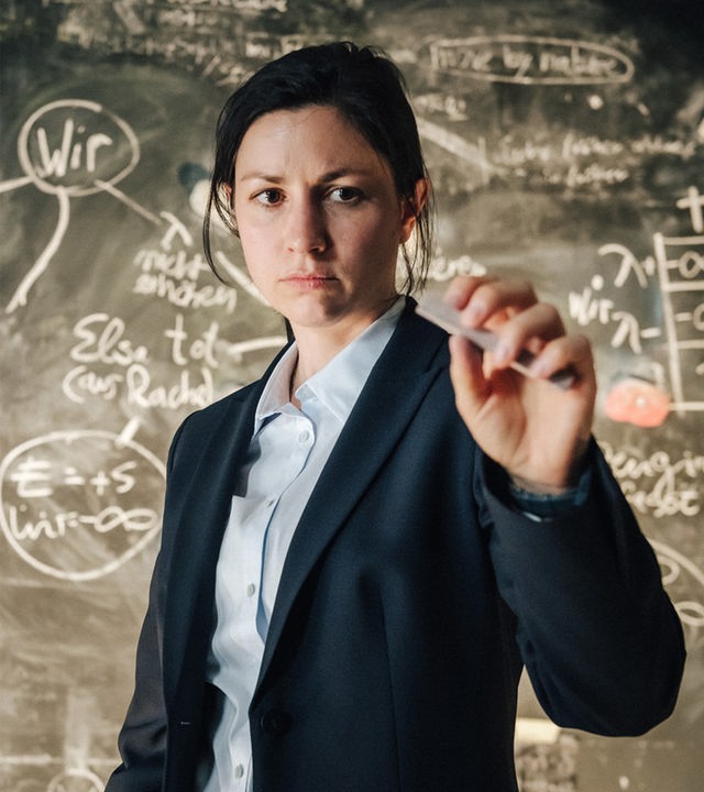 Vera Schröder (Eva Meckbach) steht vor einer Tafel mit mathematischen Formeln und schaut konzentriert auf ein stück Kreide was sie auf Augenhöhe vor sich hält.