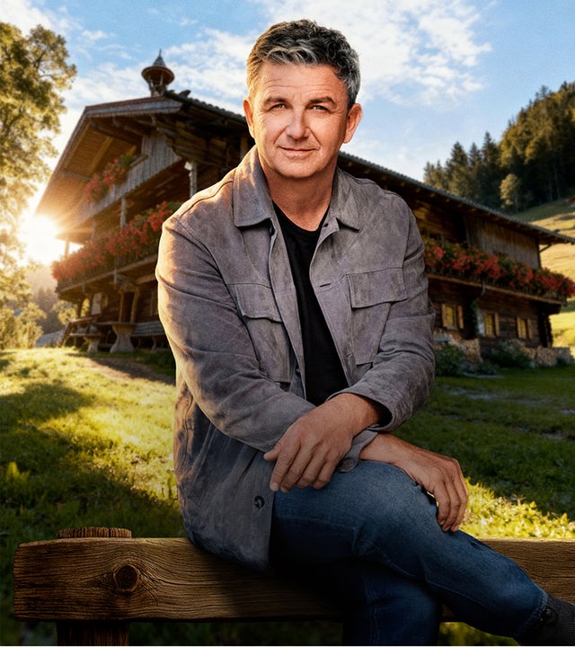 Der Bergdoktor – Dr. Martin Gruber (Hans Sigl) sitzt auf der Rückenlehne einer Holzbank. Er wird von der tiefstehenden Abensonne beleuchtet. Im Hintergrund ist seine Hütte in mitten einer verträumten Berglandschaft zu sehen. Er hat sein rechtes Bein lässig über das linke geschlagen und seine Oberarme auf den Oberschenkeln abgelegt.