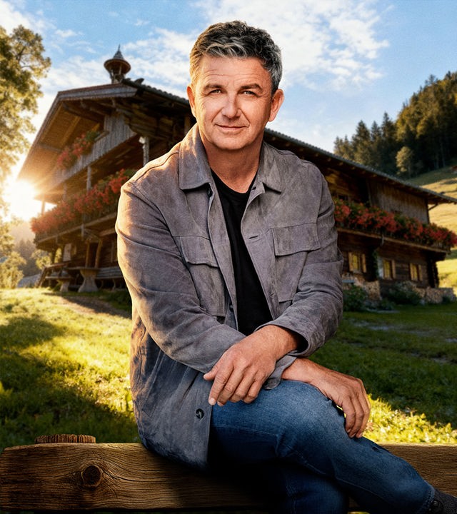 Der Bergdoktor – Dr. Martin Gruber (Hans Sigl) sitzt auf der Rückenlehne einer Holzbank. Er wird von der tiefstehenden Abensonne beleuchtet. Im Hintergrund ist seine Hütte in mitten einer verträumten Berglandschaft zu sehen. Er hat sein rechtes Bein lässig über das linke geschlagen und seine Oberarme auf den Oberschenkeln abgelegt.