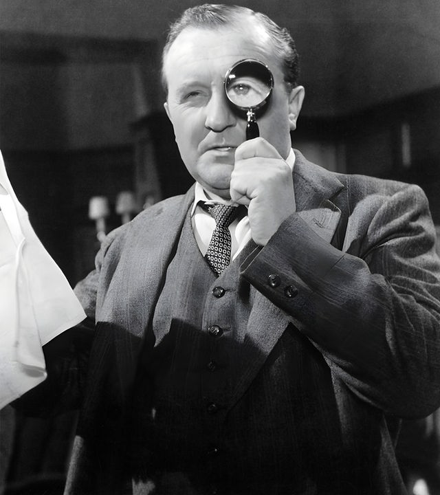 Spielszene in Schwarz-Weiß: Ein Mann im Anzug hält sich eine Lupe vor das linke Auge, in der rechten Hand hält er ein Taschentuch. Im Hintergrund beobachtet ihn ein bärtiger Mann in Hemd und Jacke.