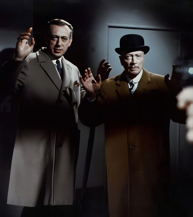 Inspektor Perkins (Horst Tappert) und Sir Arthur (Hubert von Meyerinck) stehen mit erhobenen Händen nebeneinander in einem düsteren Raum und blicken ernst, während eine Waffe bedrohlich auf sie gerichtet ist.