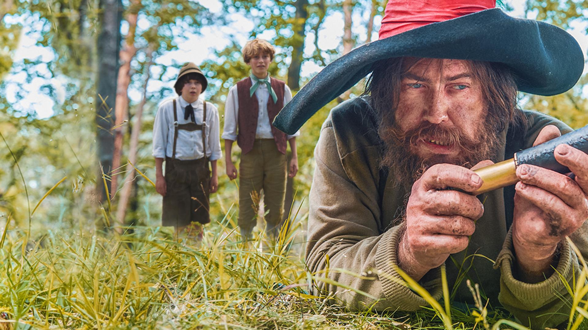 Die Jungen Kasperl und Seppel haben sich von hinten an den Räuber Hotzenplotz herangeschlichen und sehen erstaunt aus, ihn gefunden zu haben. Der Räuber, mit dreckiger Kleidung und großem Schlapphut, liegt im Vordergrund auf dem Boden und betrachtet das Messer in seiner Hand.