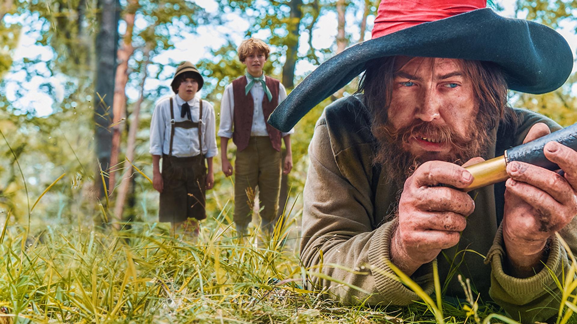 Die Jungen Kasperl und Seppel haben sich von hinten an den Räuber Hotzenplotz herangeschlichen und sehen erstaunt aus, ihn gefunden zu haben. Der Räuber, mit dreckiger Kleidung und großem Schlapphut, liegt im Vordergrund auf dem Boden und betrachtet das Messer in seiner Hand.