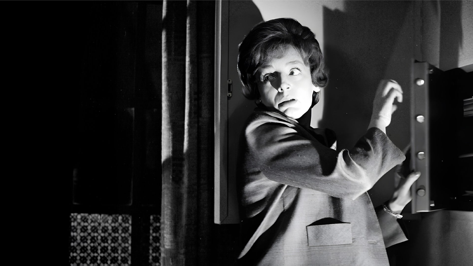 Eine Frau steht vor einem halb geöffneten Wandsafe im Halbschatten, schlaglichtartig wird ihr Gesicht von einer einzelnen Lichtquelle erhellt. Vorsichtig blickt sie über ihre Schulter.