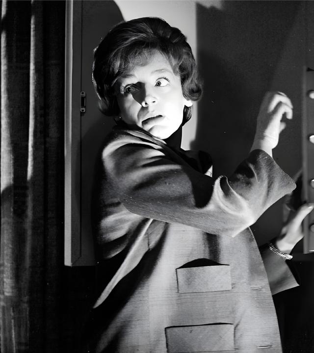 Eine Frau steht vor einem halb geöffneten Wandsafe im Halbschatten, schlaglichtartig wird ihr Gesicht von einer einzelnen Lichtquelle erhellt. Vorsichtig blickt sie über ihre Schulter.