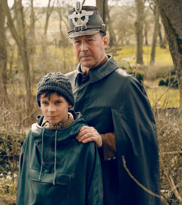 "Deutschstunde": Jens Ole Jepsen (Ulrich Noethen) und Siggi Jepsen (Levi Eisenblätter) stehen nebeneinander und schauen in die Kamera. Jens Ole hat seine Hände auf die Schultern seines Sohnes gelegt.