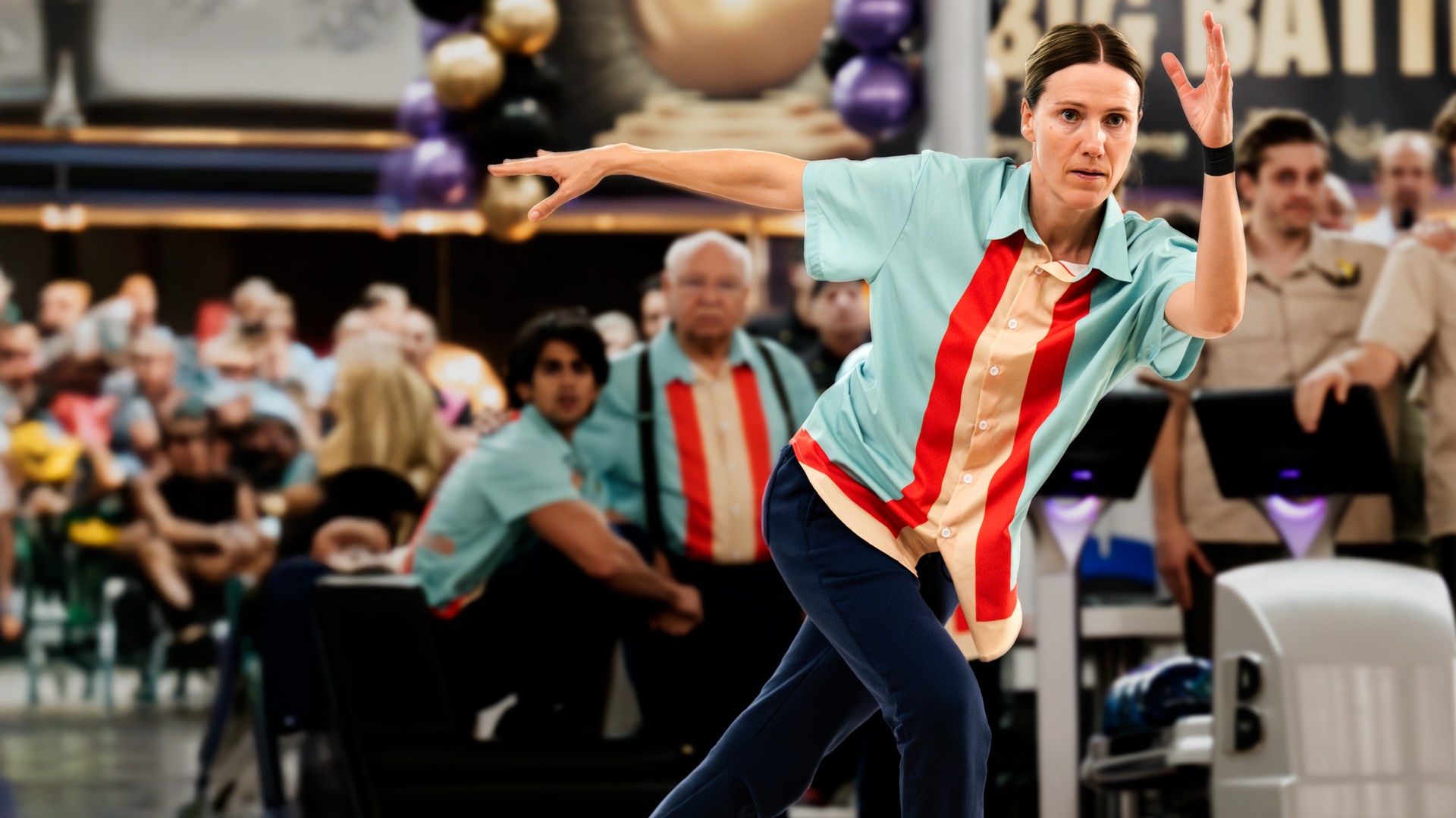 Karo (Bettina Lamprecht) rollt ihre Bowlingkugel mit angespanntem Blick auf die Bowlingbahn. Hinter ihr steht ihr Team, daneben das gegnerische Team (Komparsen).