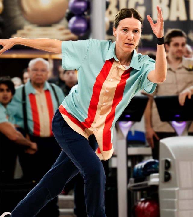 Karo (Bettina Lamprecht) rollt ihre Bowlingkugel mit angespanntem Blick auf die Bowlingbahn. Hinter ihr steht ihr Team, daneben das gegnerische Team (Komparsen).