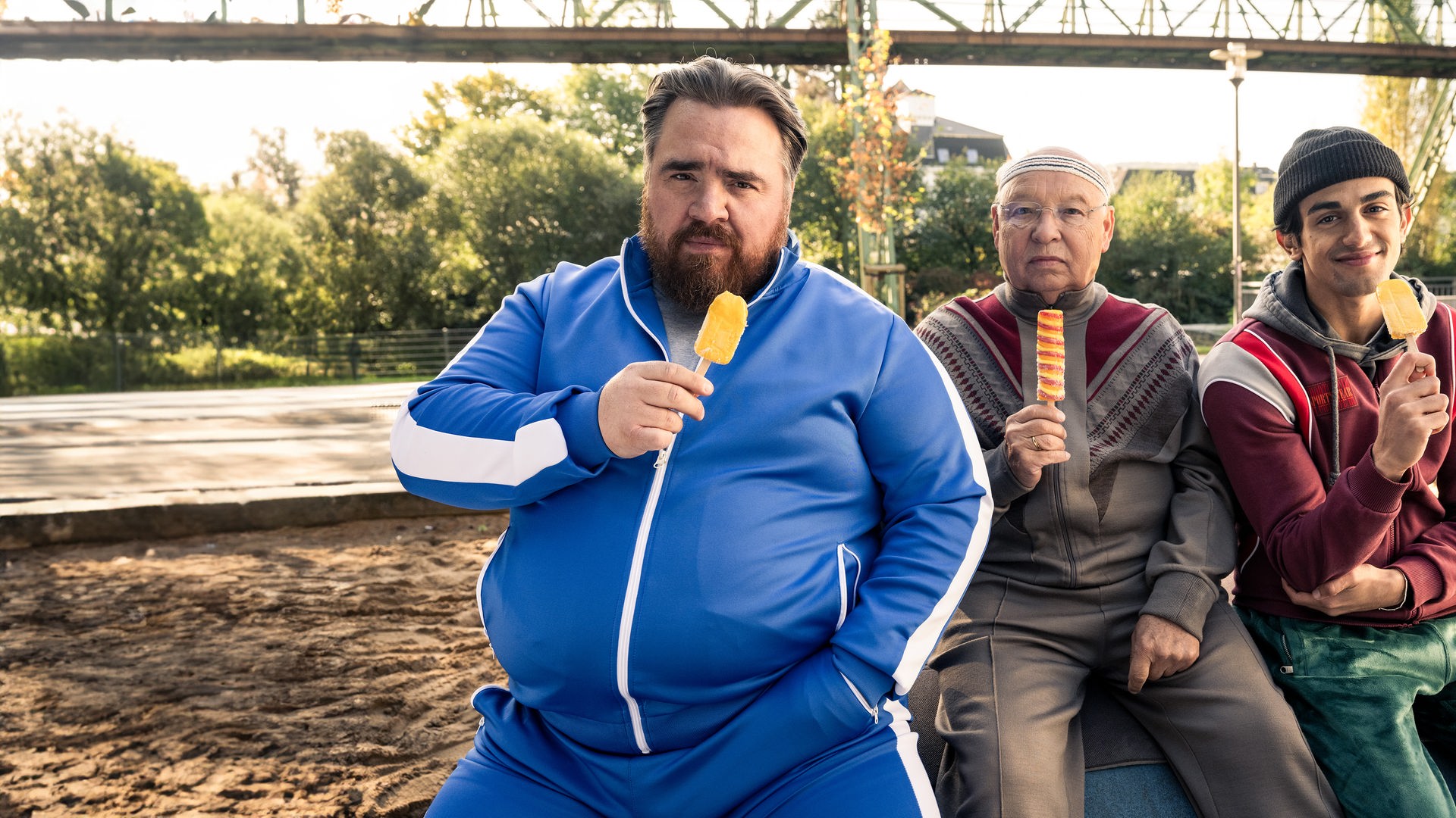 "Die Abräumer": Axel (Tristan Seith), Freddy (Hans-Joachim Heist) und Djamal (Mido Kotaini) sitzen nebeneinander im Freien, schauen in die Kamera und halten alle ein Eis am Stiehl in der Hand.