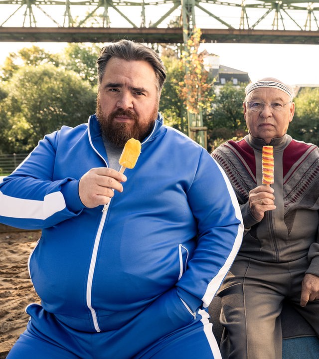 "Die Abräumer": Axel (Tristan Seith), Freddy (Hans-Joachim Heist) und Djamal (Mido Kotaini) sitzen nebeneinander im Freien, schauen in die Kamera und halten alle ein Eis am Stiehl in der Hand.