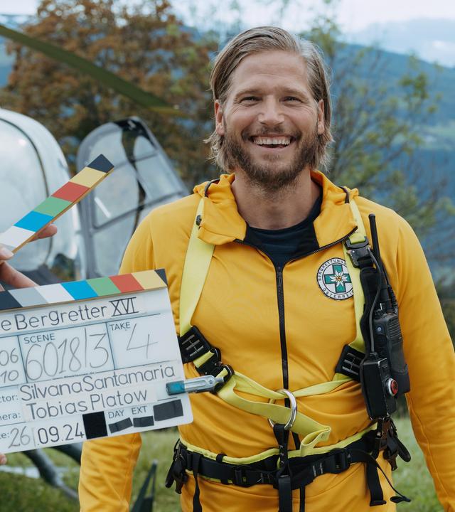Sebastian Ströbel aus der Serie "Die Bergretter" steht vor einem Rettungs-Helikopter. Vor ihm sieht man eine Filmklappe, denn das Foto entstand während der Dreharbeiten. Die Doku Backstage schaut hinter die Kulissen der Serie.