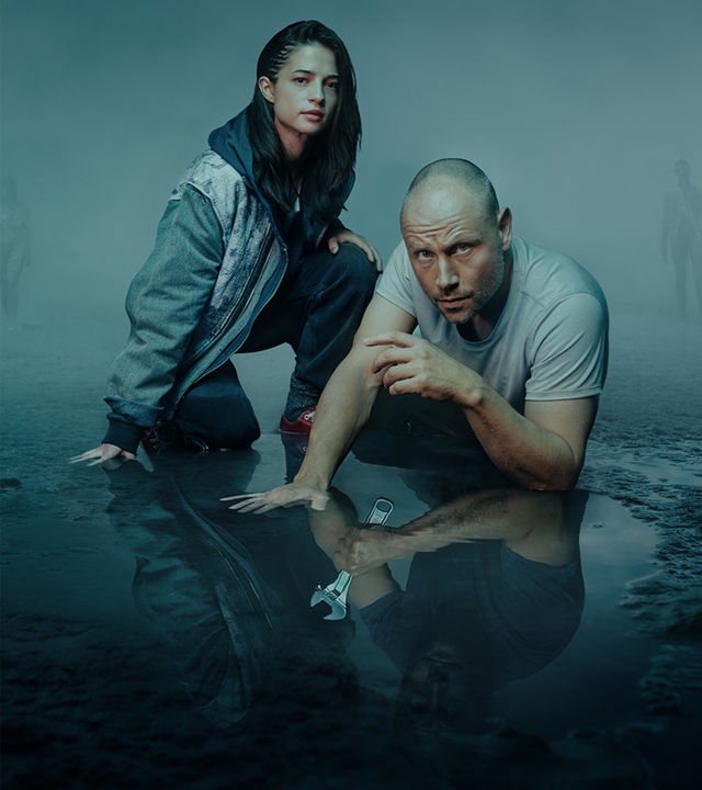 Die Düsteren: Soma (Lisa-Marie Koroll) und Ben Sandmann (Max Riemelt) knien in einer nebligen, düsteren Moorlandschaft auf dem feuchten Boden. Sie blicken mit ernstem Ausdruck direkt in die Kamera, während sich ihre Gestalten im spiegelnden Wasser vor ihnen abzeichnen.