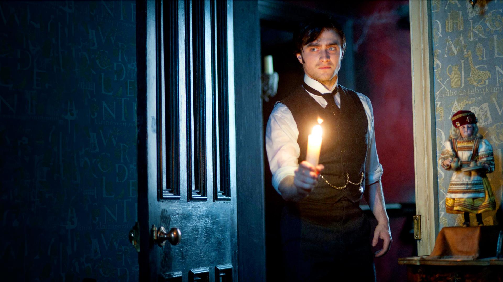 Arthur Kipps (Daniel Radcliffe) steht in einem Türrahmen und hält eine brennende Kerze in der rechten Hand. Er trägt ein weißes Hemd, mit schwarzer Krawatte und Weste. An der Weste ist eine goldene Kette befestigt, die zu einer Taschenuhr gehört.