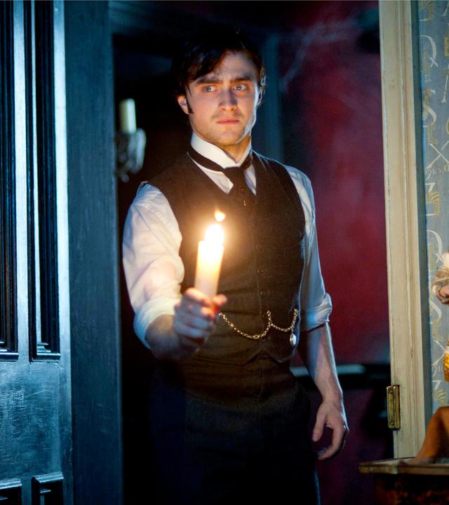 Arthur Kipps (Daniel Radcliffe) steht in einem Türrahmen und hält eine brennende Kerze in der rechten Hand. Er trägt ein weißes Hemd, mit schwarzer Krawatte und Weste. An der Weste ist eine goldene Kette befestigt, die zu einer Taschenuhr gehört.