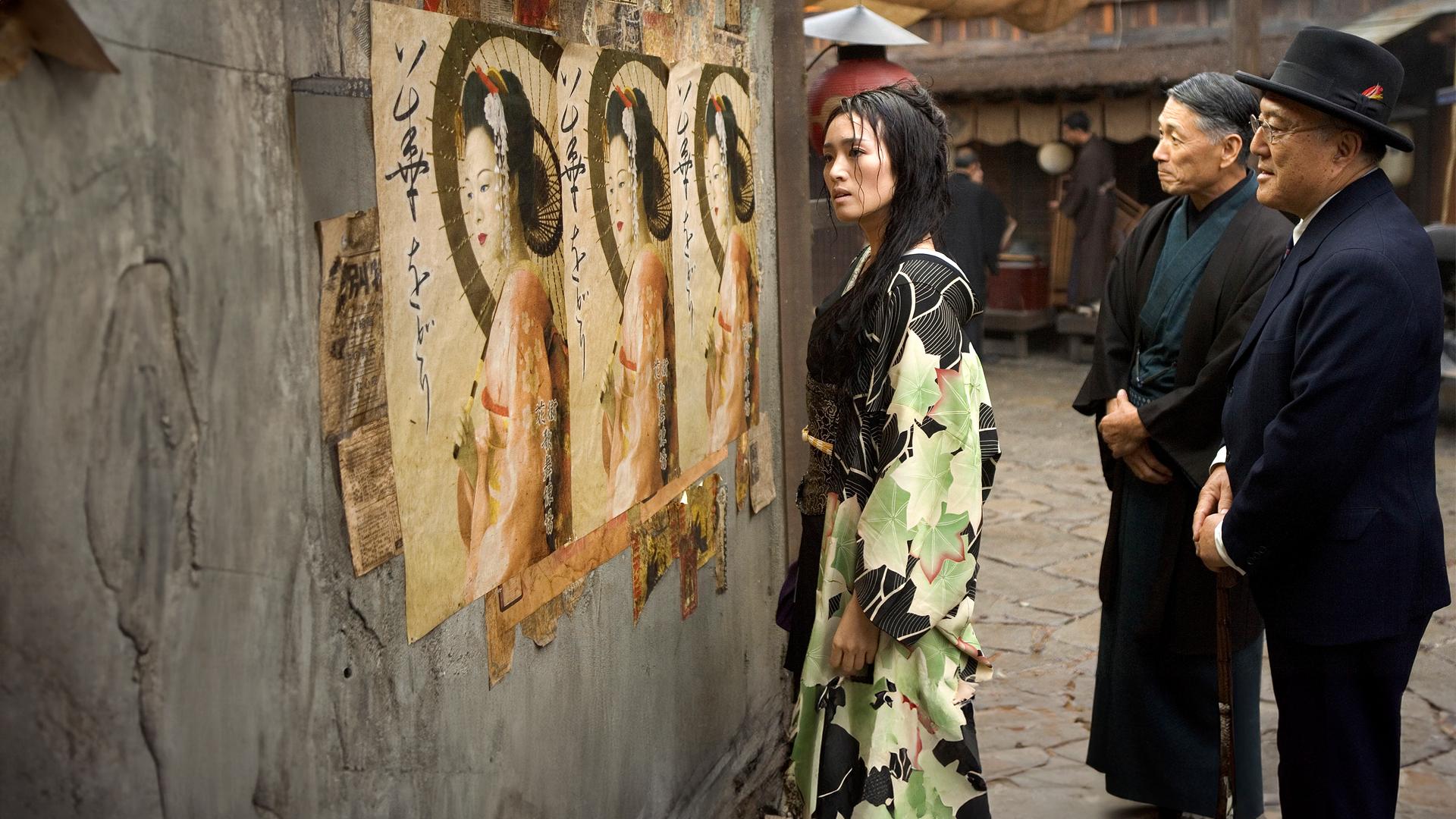  Geisha Hatsumomo (Gong Li) steht auf der Straße und schaut sich Plakate an, die eine Geisha zeigen. Hinter ihr stehen zwei Männer (Darsteller nicht bekannt), die ebenfalls auf die Plakate schauen.