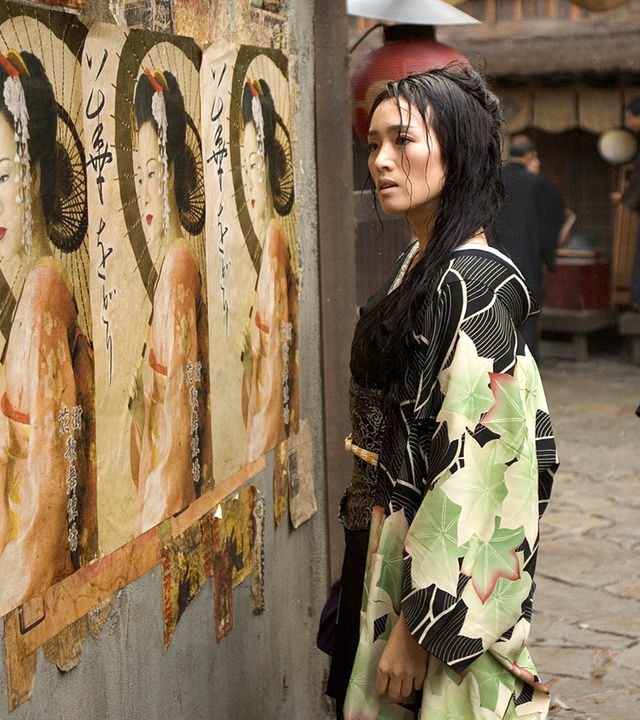  Geisha Hatsumomo (Gong Li) steht auf der Straße und schaut sich Plakate an, die eine Geisha zeigen. Hinter ihr stehen zwei Männer (Darsteller nicht bekannt), die ebenfalls auf die Plakate schauen.