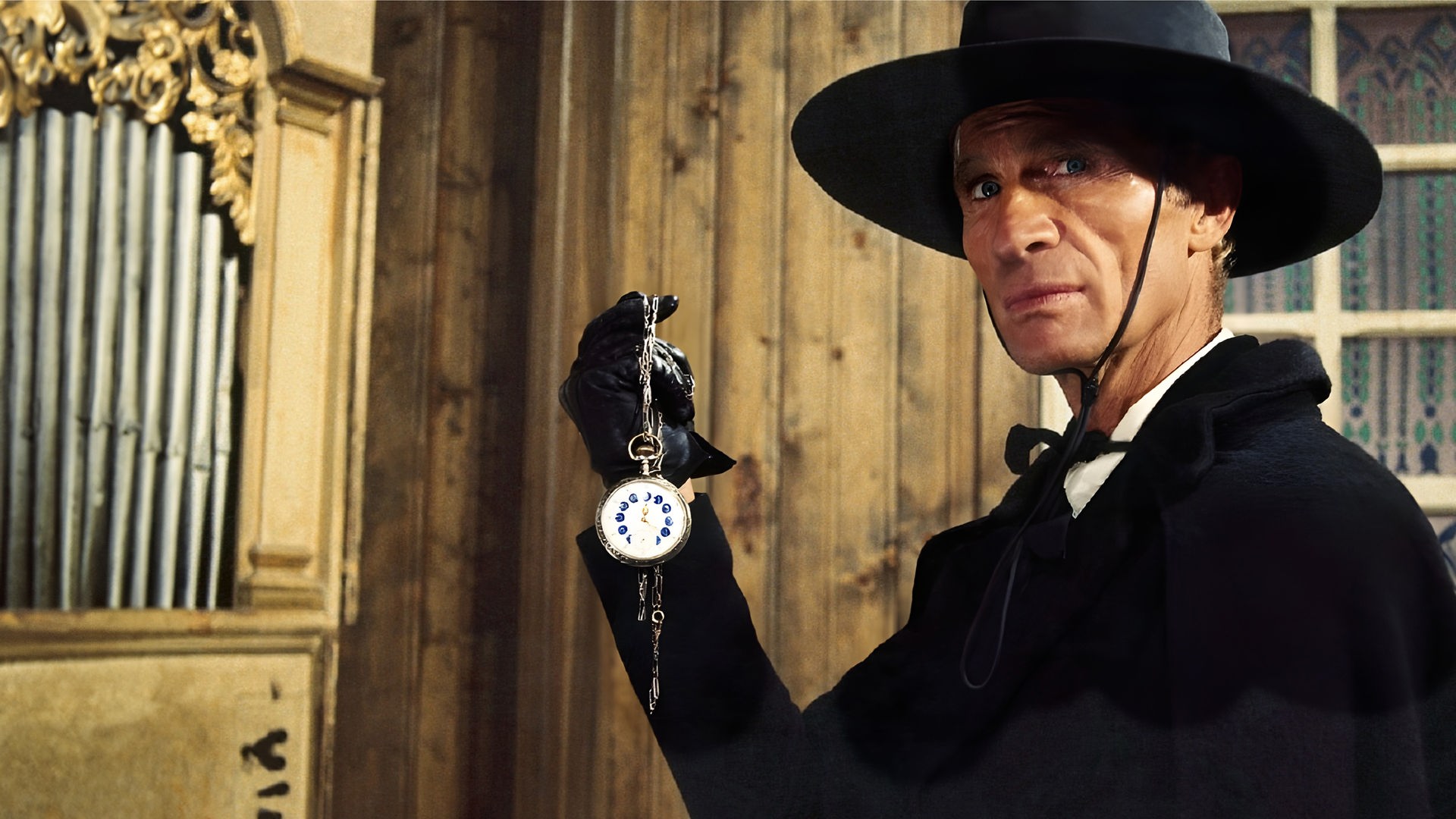 Burton (Gordon Mitchell) trägt einen schwarzen Mantel, einen breitkrempigen Hut und schwarze Handschuhe, während er eine Taschenuhr an einer Kette in die Höhe hält und mit ernstem Ausdruck geradeaus blickt. Hinter ihm sind holzvertäfelte Wände sowie ein vergittertes Fenster zu sehen.