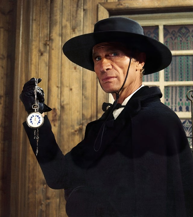 Burton (Gordon Mitchell) trägt einen schwarzen Mantel, einen breitkrempigen Hut und schwarze Handschuhe, während er eine Taschenuhr an einer Kette in die Höhe hält und mit ernstem Ausdruck geradeaus blickt. Hinter ihm sind holzvertäfelte Wände sowie ein vergittertes Fenster zu sehen.