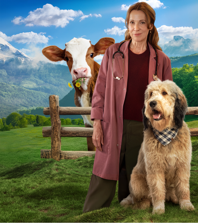 Johanna Maiwald (Doris Schretzmayer), Tierärztin in Maria Alm, steht vor einer grünen Bergkulisse mit eisbedeckten Spitzen. Sie trägt ein Stethoskop um den Hals. Vor ihr sitzt ein großer langhaariger Hund mit einem Stofftuch um den Hals. Hinter ihr schaut neugierig der Kopf eines Kalbs hervor. es hat ein paar Löwenzahnblüten im Maul.