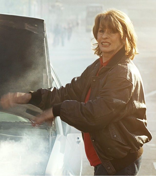 Taxifahrerin Gerda (gespielt von Senta Berger) steht vor der offenen Kühlerhaube ihres Taxis. Aus dem Motorraum steigt Rauch hervor.