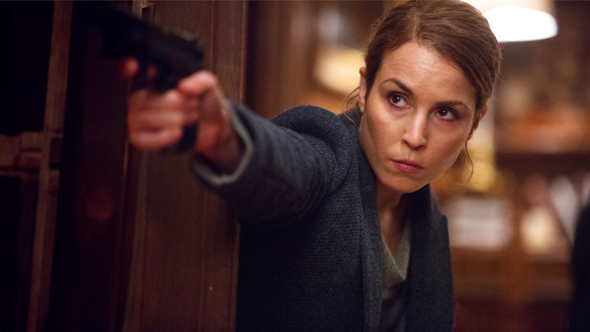 Alice Racine (Noomi Rapace) steht neben einem Türrahmen und zielt mit ihrer Pistole.