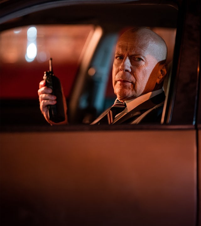Die Spur des Killers – Midnight in the Switchgrass": FBI-Agent Karl Helter (Bruce Willis) in Anzug, Krawatte und mit einem FBI-Abzeichen um den Hals, sowie seine Kollegin Rebecca Lombardi (Megan Fox) in Zivil stehen im Freien vor einem von der Polizei abgesperrten Bereich. Im Hintergrund stehen diverse Autos.