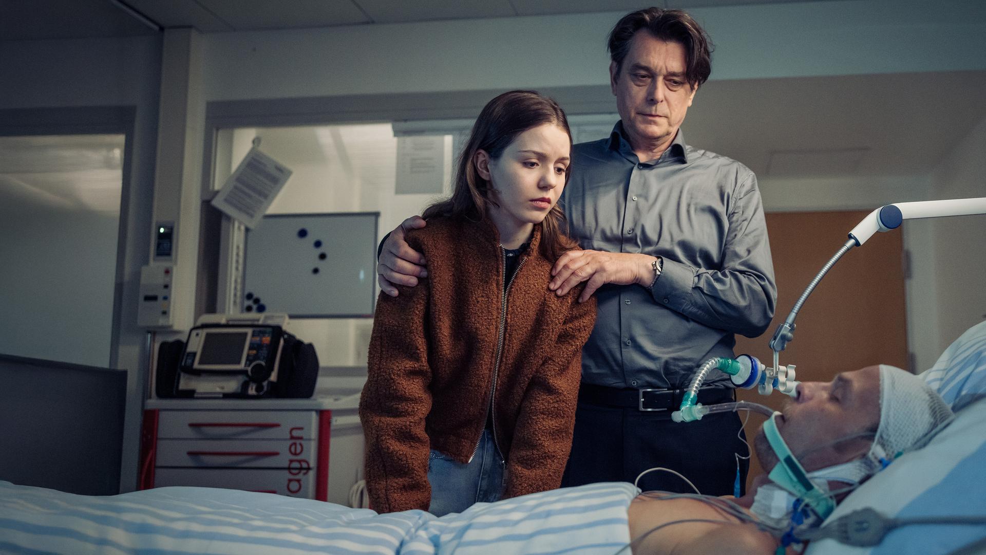 "Die Toten vom Bodensee - Im Schattenreich": Luna Oberländer (Fiona Katharina Neumeier) und Thomas Komlatschek (Hary Prinz) stehen bei Micha Oberländer (Matthias Koeberlin) am Krankenbett.