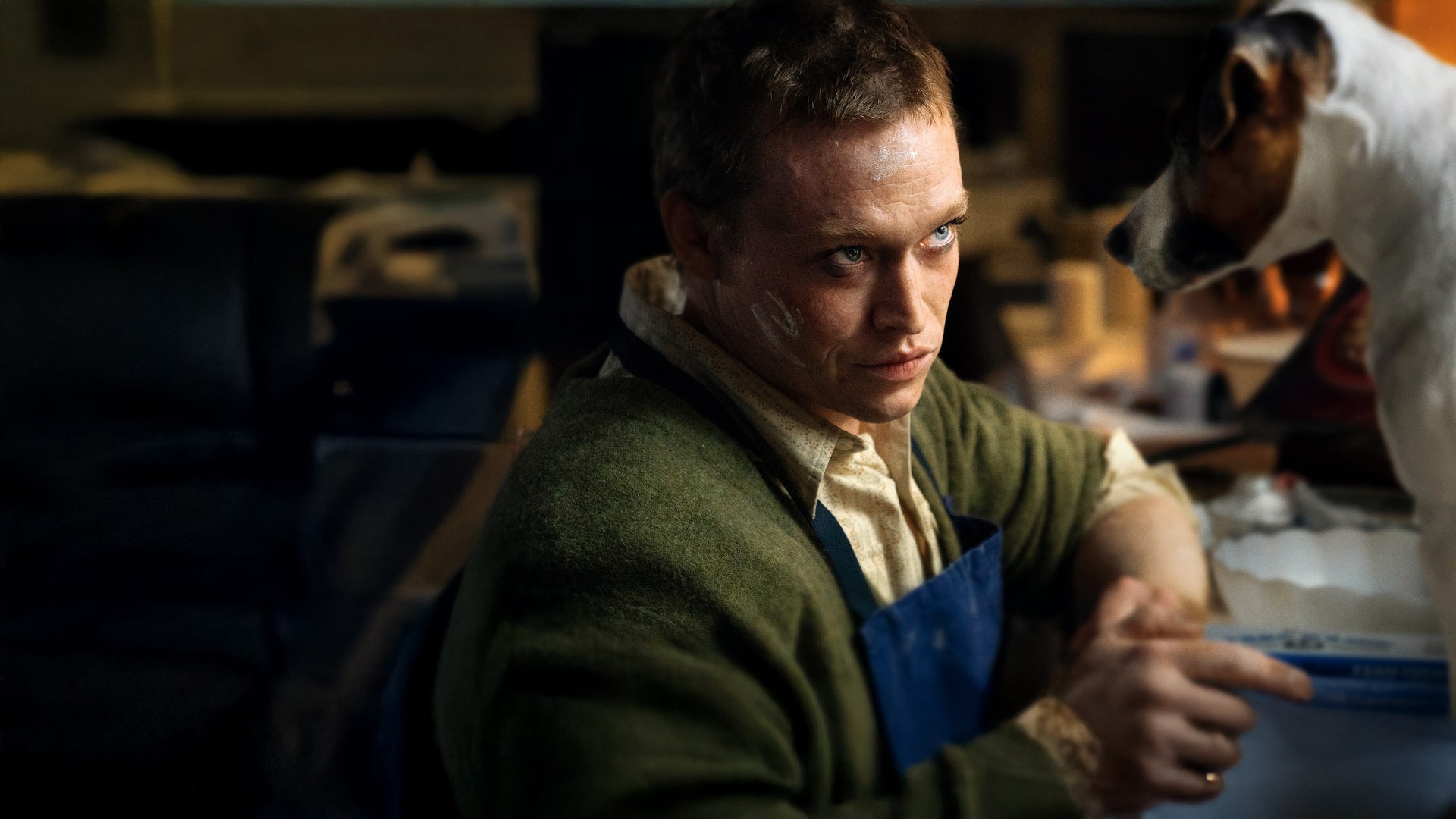 "DogMan": Doug Munrow (Caleb Landry Jones) sitzt mit einer blauen Schürze vor einer Anrichte mit Utensilien zum Kuchenbacken. Von ihm sitzt ein kleiner Hund auf der Anrichte, beide schauen sich intensiv an.