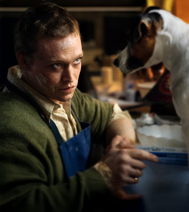 "DogMan": Doug Munrow (Caleb Landry Jones) sitzt mit einer blauen Schürze vor einer Anrichte mit Utensilien zum Kuchenbacken. Von ihm sitzt ein kleiner Hund auf der Anrichte, beide schauen sich intensiv an.