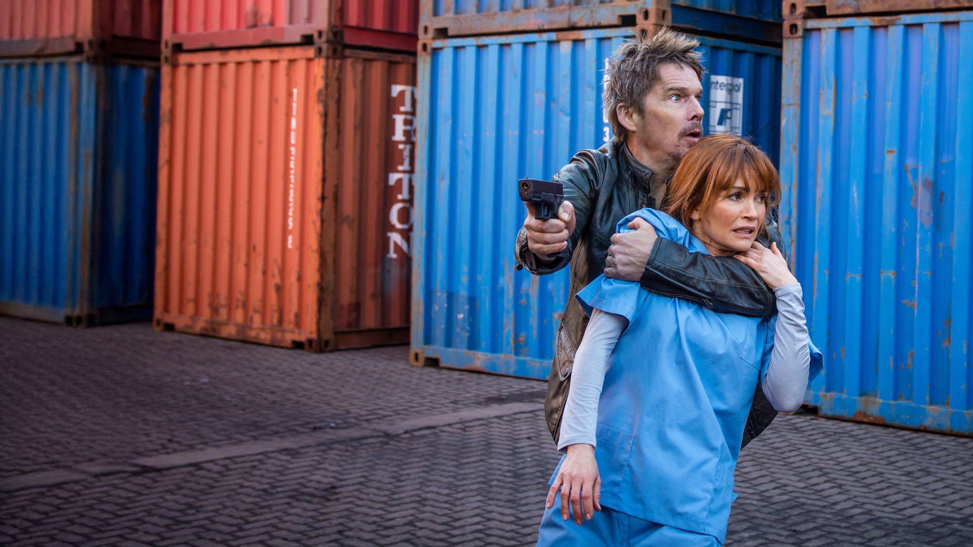ravis Conrad (Ethan Hawke) hat Dr. Helen (Nathalie Boltt) als Geisel genommen, um aus dem Labor zu fliehen. "Du lebst noch 24 Stunden": Travis Conrad (Ethan Hawke) hat eine Pistole in der Hand und umklammert Dr. Helen (Nathalie Boltt). Im Hintergrund sind mehrere große Container zu sehen.