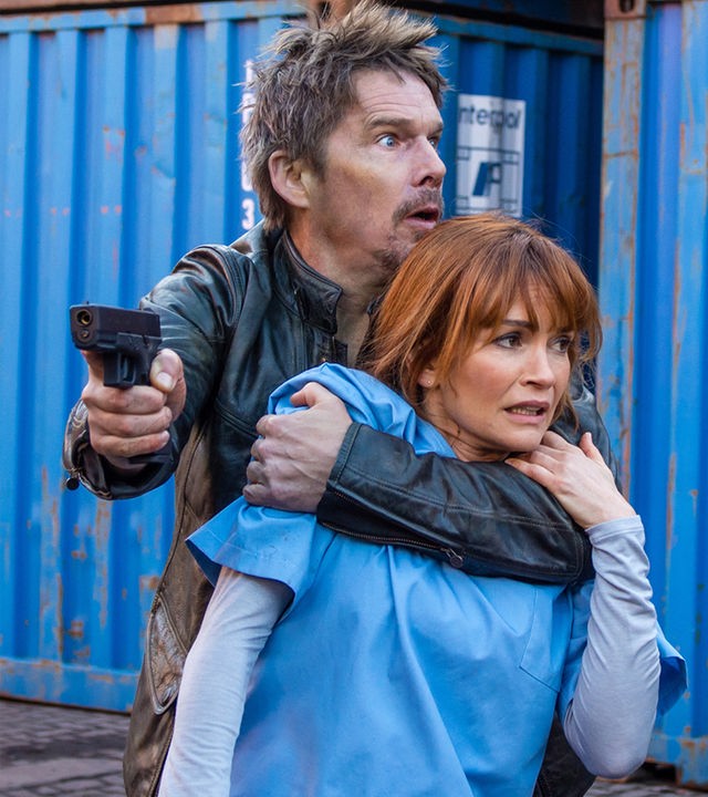 ravis Conrad (Ethan Hawke) hat Dr. Helen (Nathalie Boltt) als Geisel genommen, um aus dem Labor zu fliehen. "Du lebst noch 24 Stunden": Travis Conrad (Ethan Hawke) hat eine Pistole in der Hand und umklammert Dr. Helen (Nathalie Boltt). Im Hintergrund sind mehrere große Container zu sehen.
